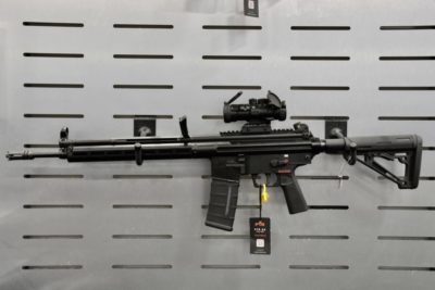 A black, tactical rifle hanging on a grey wall. The rifle has a scope on it with flip-up caps.