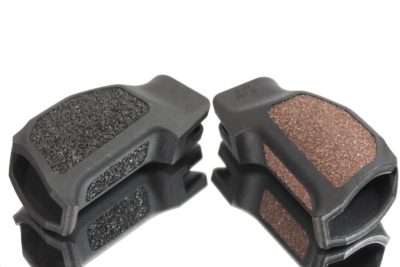 Two AR-style grips made of black plastic are laying on a mirror surface. One grip has black, rubber accents while the other has brown, sandpaper accents.