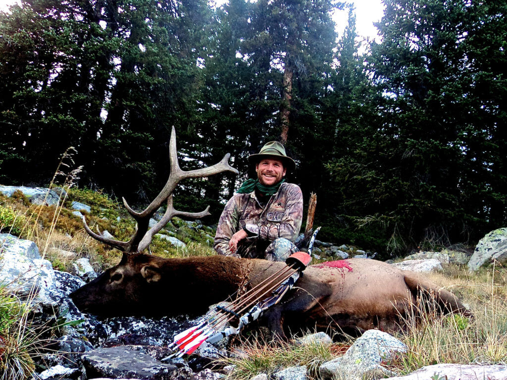 Hunter posing with his elk, longbow, and quiver in a forest clearing