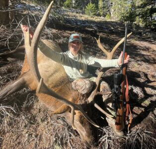 A happy hunter poses with her trophy bull elk and gun.