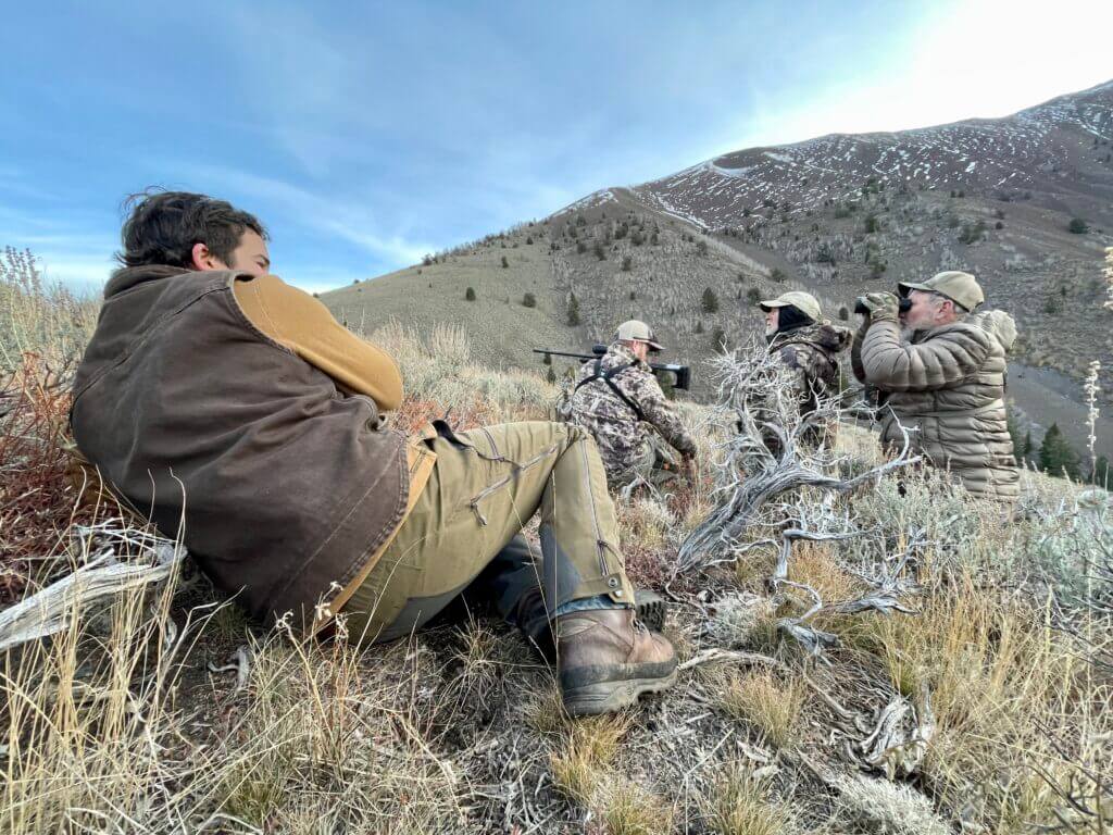 Hunters look through binoculars at their prey while sitting on a sagebrush hillside.