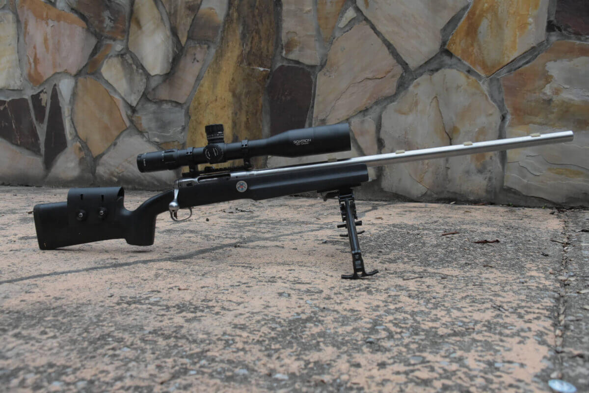 A rifle rests on a bipod with a stone background.