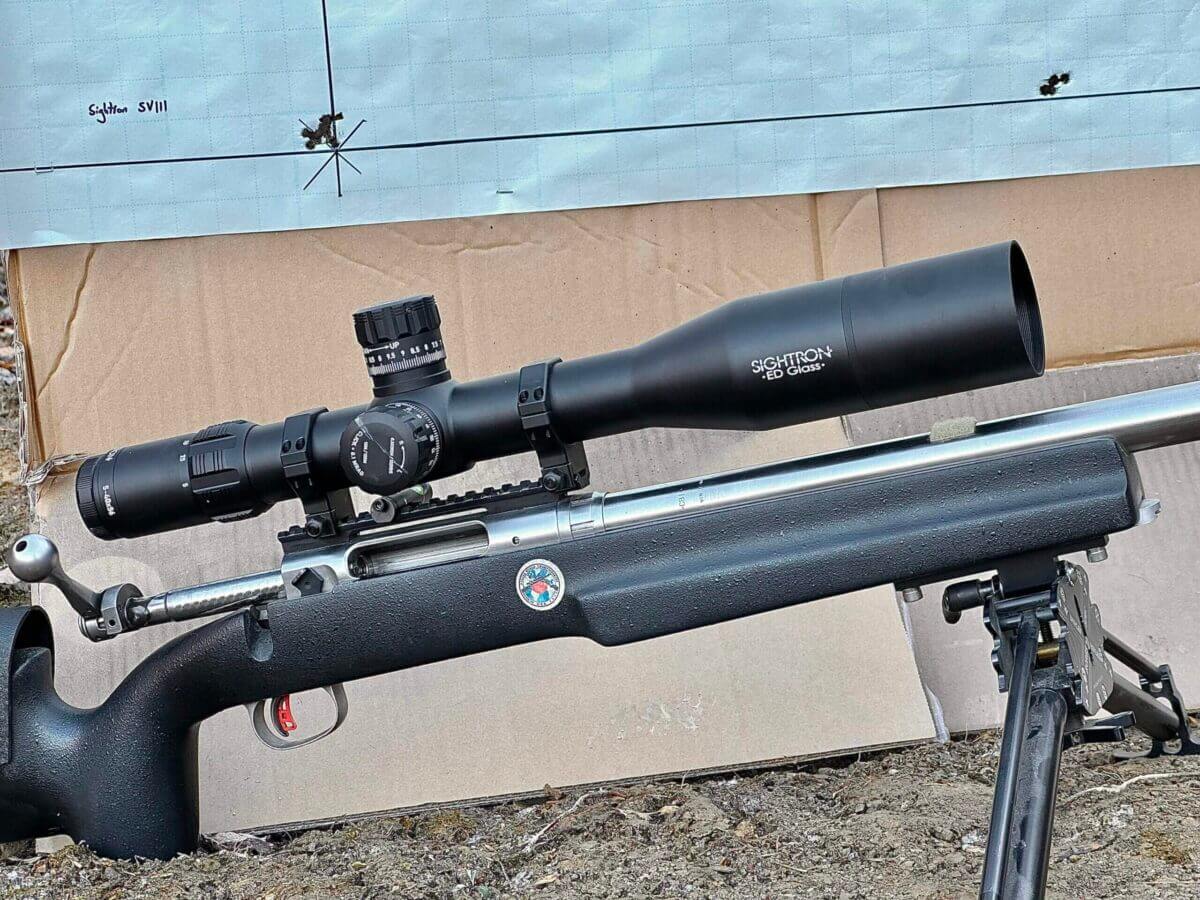 A target with bullet holes in it sits behind a target rifle with a large optic on it.