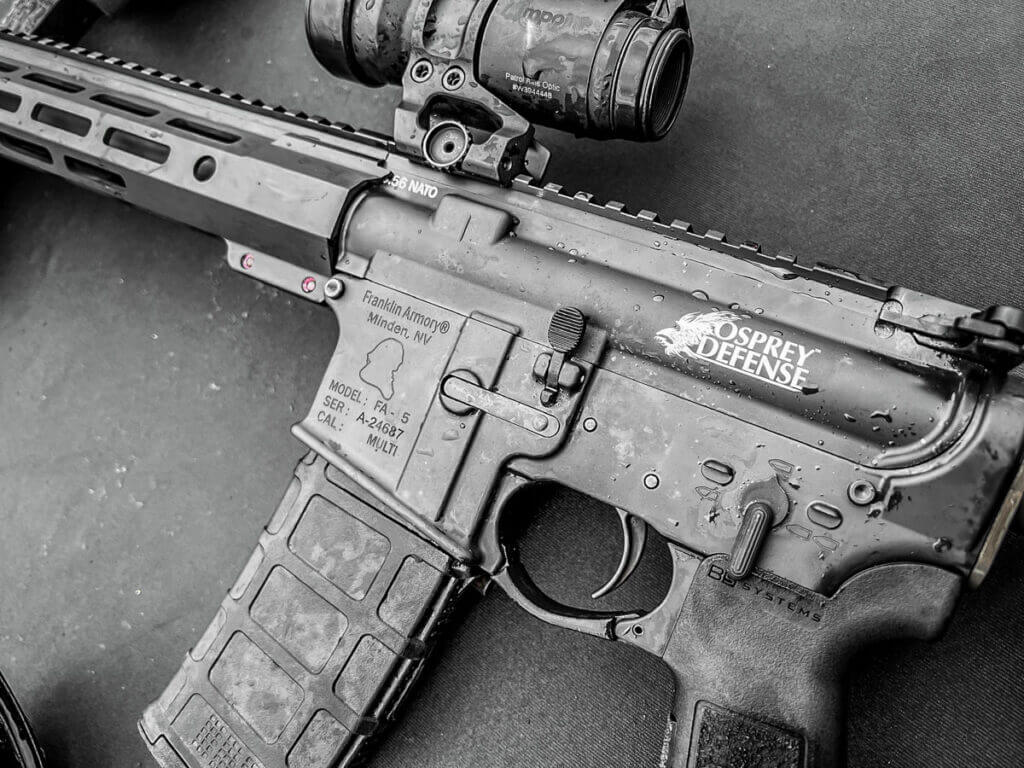 An AR-style rifle is laid on a black table. Both are covered in rain.