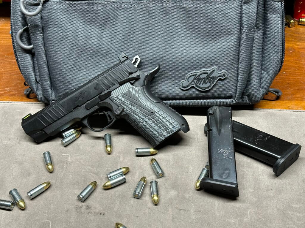 A black Kimber KDS9C handgun leans up against a soft carry-case with bullets scattered on the table next to them.