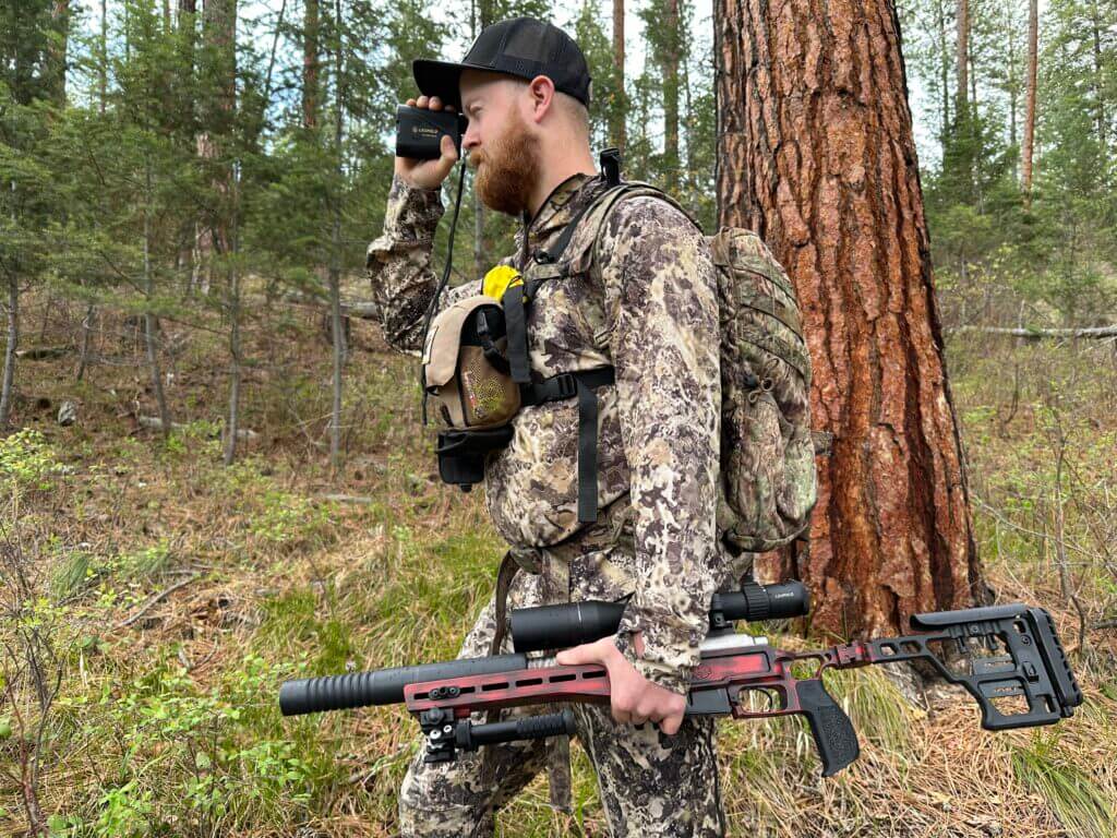 A man in camouflage is standing in the woods with a rifle and the RX-5000 in his hand.