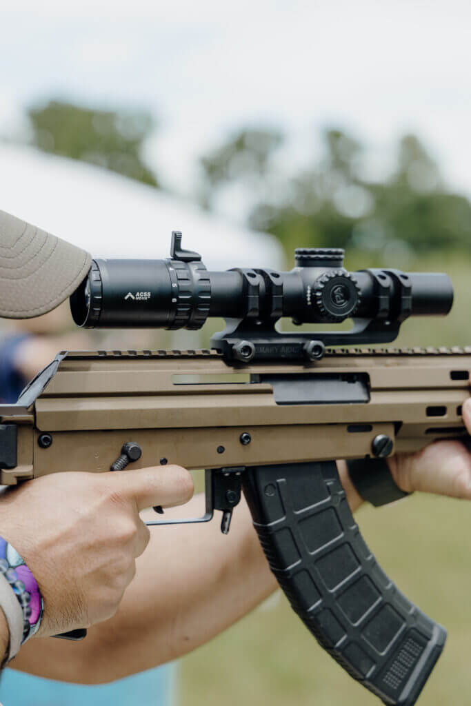 Shooter aiming down a scope mounted on a rifle out under the trees