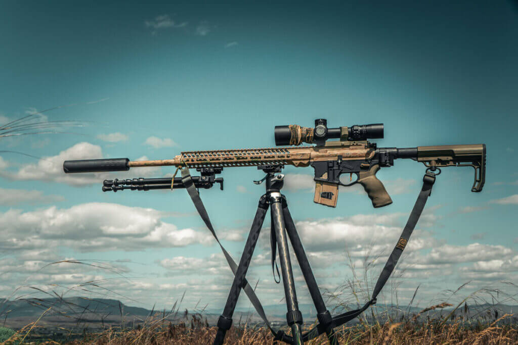 A beige rifle with a scope mounted to a tripod in front of a vibrant blue sky