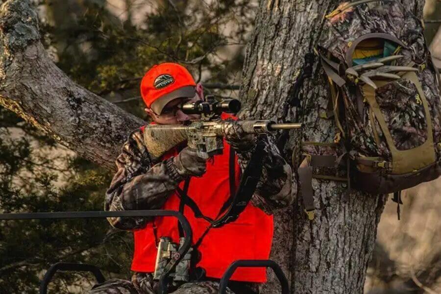 A hunter with an orange vest in a stand holding a rifle.