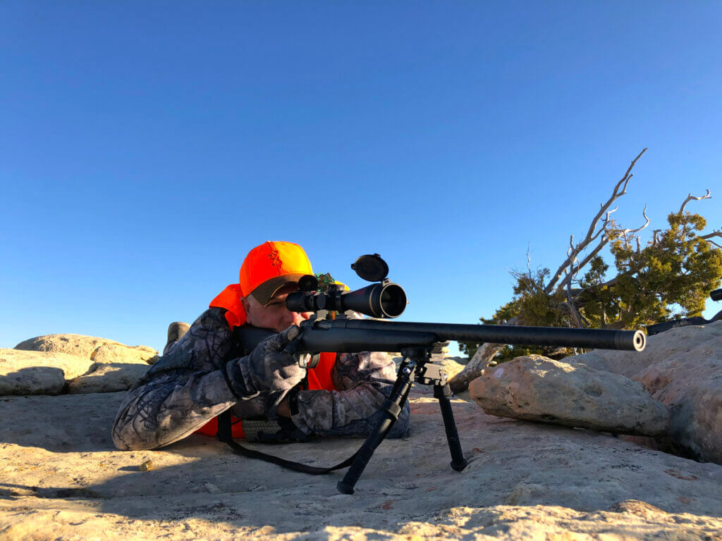 hunter prone behind his rifle on a sandy rock