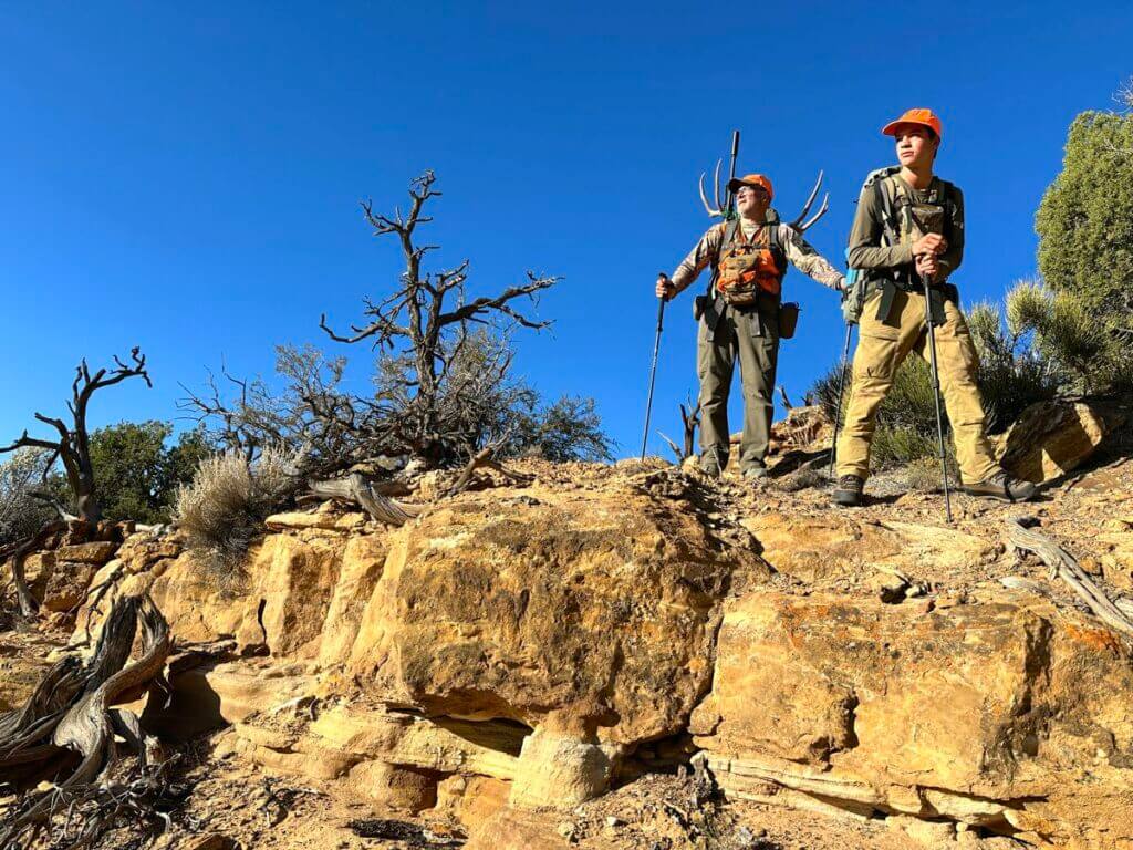 Two hunters in camo and orange packing a mule deer buck out of the desert.