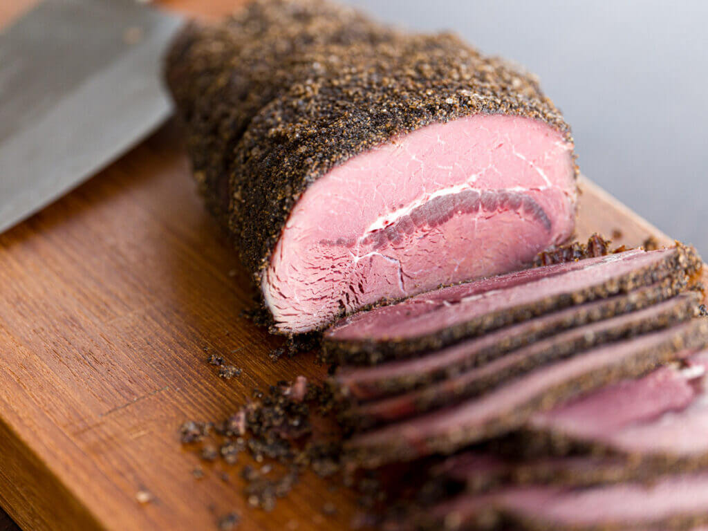 Picture of elk pastrami sliced across the grain on a cutting board with a knife in the background.