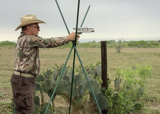man wearing camo shirt and cowboy hat standing outside at a tripod holding and aiming a pistol