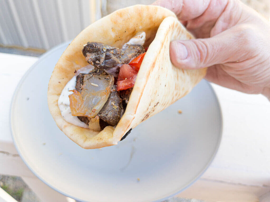 The pita full of fixings ready to eat, held in the photographer's hand over a plate on a deck railing outdoors