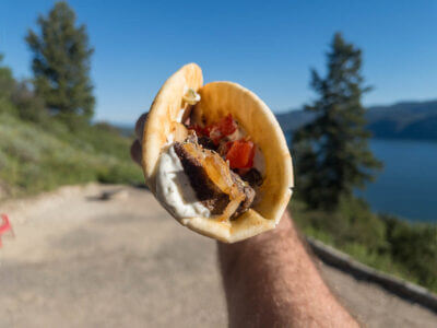 A loaded pita with meat and fixings nearly spilling out, held by the photographer's hand with a mountain lake scene in the background.