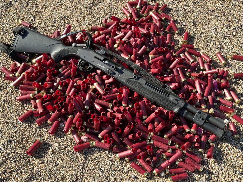 A black shotgun lays on a pile of red shotgun shells on the ground.