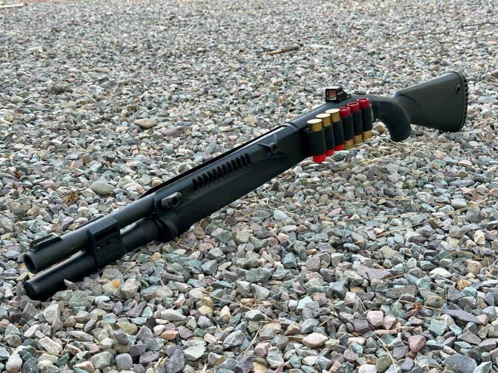 A shotgun lays in the gravel with shotgun shells strapped to its side. 