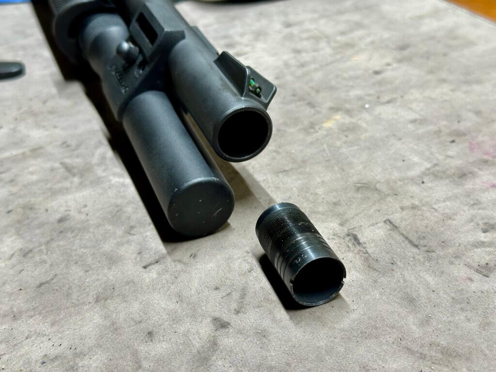 the choke tube is removed from a shotgun barrel and is being showcased on a tan mat, next to the gun. 