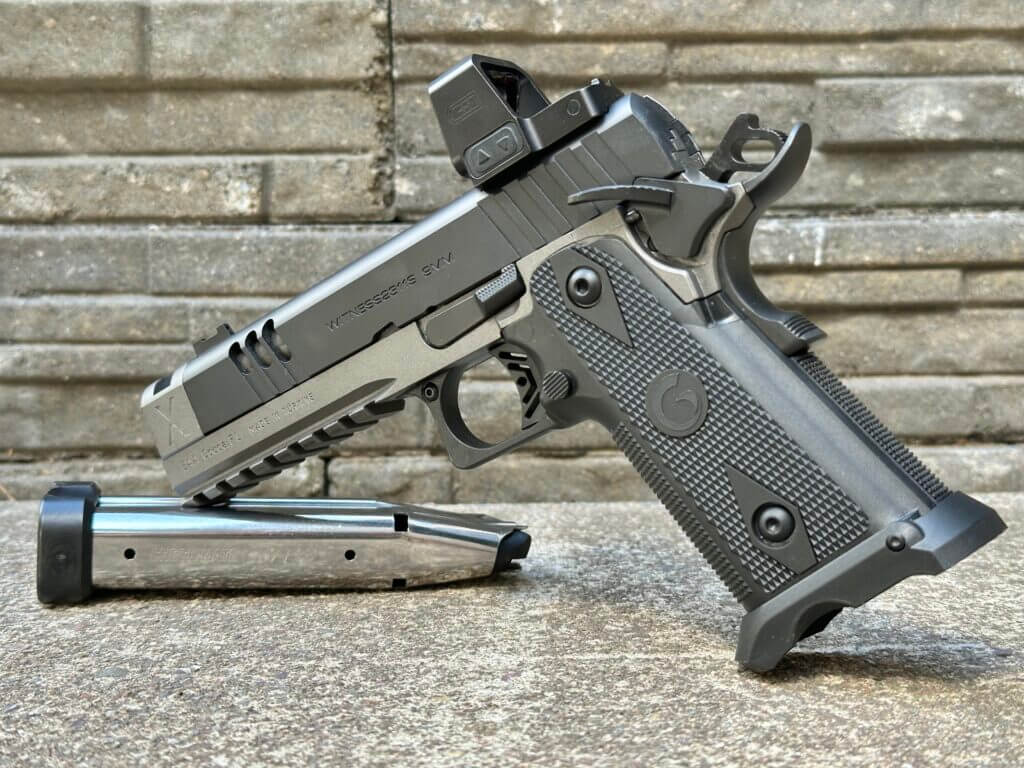 A black and silver handgun with an optic is staged by a silver magazine in front of a stone backdrop. Witness2311 Match X