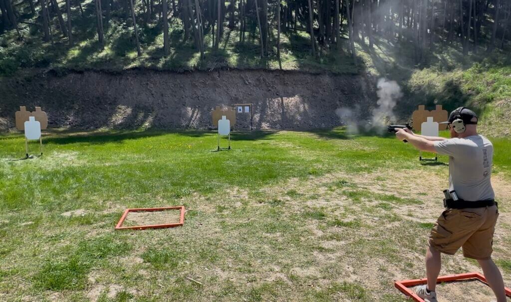 A man is shooting a gun at the gun range. An array of targets are in the background. MAX