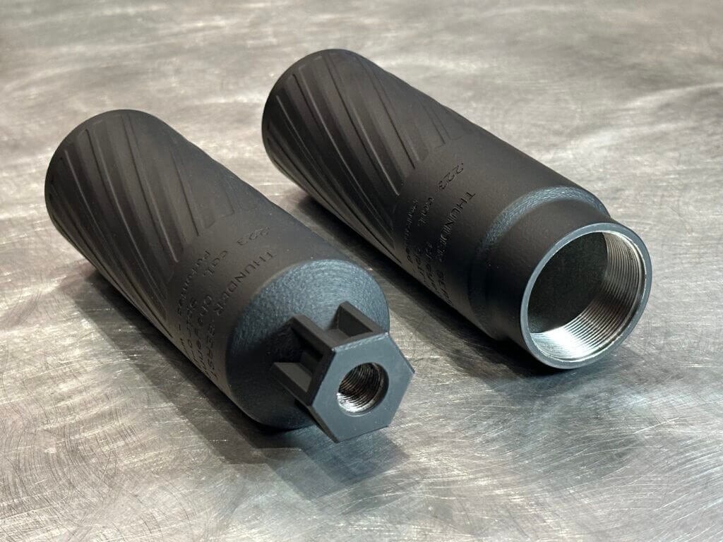 Two suppressors lay on a steel table. These black cans are faced away, with the mounting interface toward us.