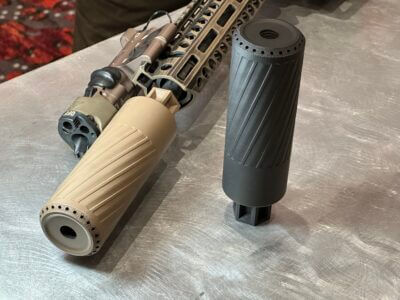 Two suppressors are being showcased. one is black and sitting on a steel tabletop and the other is tan and is mounted to a gun.