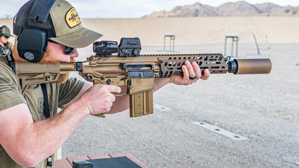 A closeup view of a man holding the M7 rifle, ready to fire. The background is a gravel shooting range with mountains in the background.