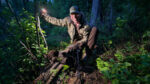 The author in the woods at near dark with a dead bear at his feet holding a cell phone flashlight up to the side for illumination on himself and the bear.