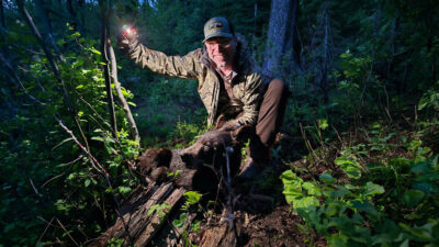 The author in the woods at near dark with a dead bear at his feet holding a cell phone flashlight up to the side for illumination on himself and the bear.