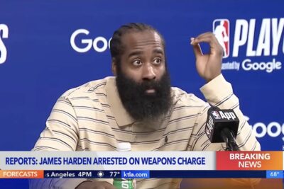 James Harden speaks during a postgame press conference while seated behind a microphone. A KTLA breaking news banner reports his arrest on a weapons charge.