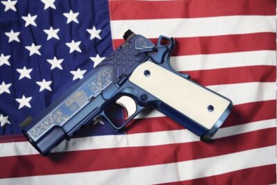 The SK Guns "250 Years of Freedom" 1911 Commander in .45 ACP is displayed atop an American flag, showcasing its sapphire PVD finish, ivory-colored grips, and patriotic engravings commemorating the United States' 250th anniversary.