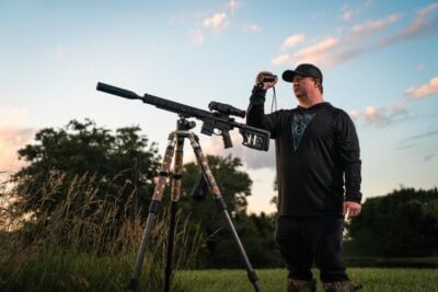 Hunter using the Athlon Cronus ATS P-MINI 250S thermal monocular while observing a field beside a suppressed rifle mounted on a tripod.