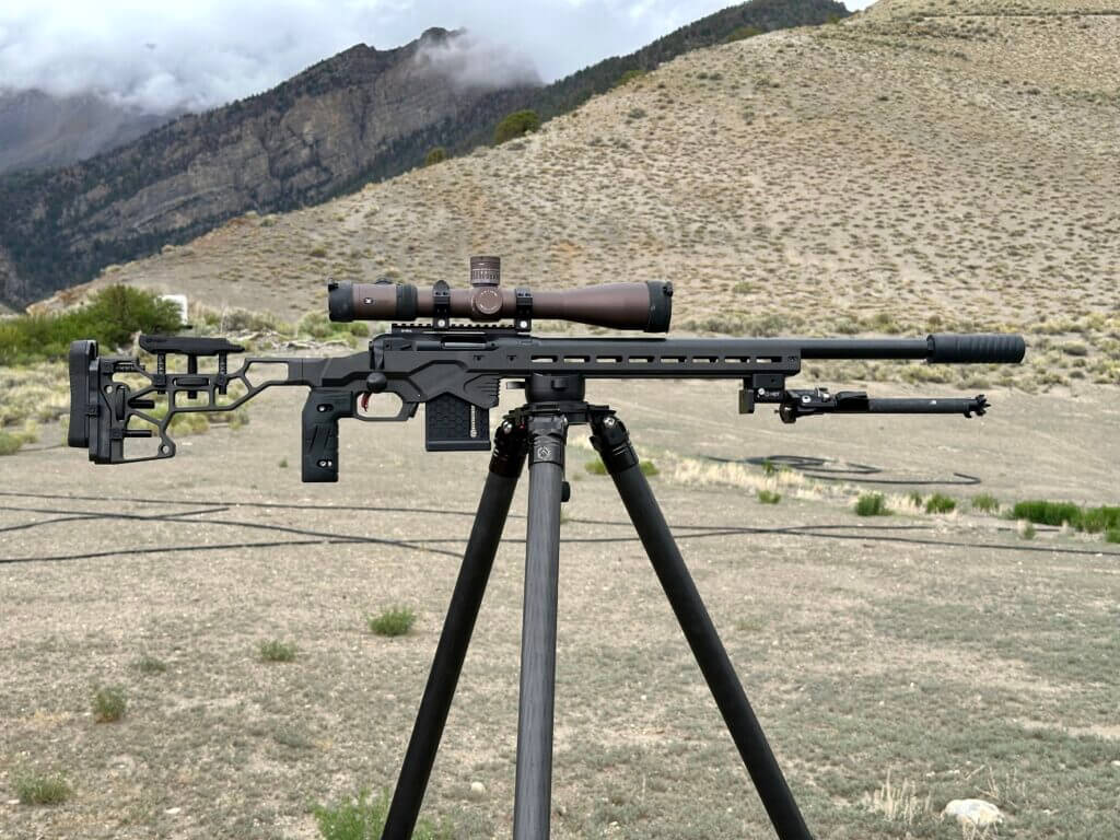A suppressed bolt action rifle on a tripod with mountains in the background.