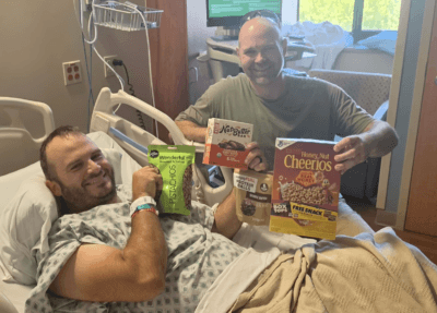 The Northwind Aerial creator smiles from a hospital bed while a friend visits with snacks after a life-changing accidental firearm discharge during a training exercise.