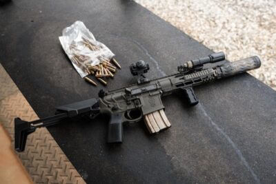 Primary Weapons Systems MK107 MOD 2-M .300 Blackout PDW with a suppressor, folding stock, red dot optic and loaded magazine resting on a shooting bench beside ammunition.
