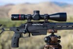 Medium close photo of the scope on a rifle mounted to a tripod with mountains in the background.