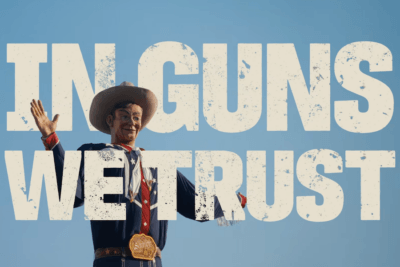 In Guns We Trust documentary title displayed over the Big Tex cowboy statue against a blue sky.