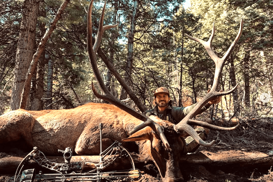 Country singer Riley Green kneels behind a large Utah bull elk harvested with a compound bow.